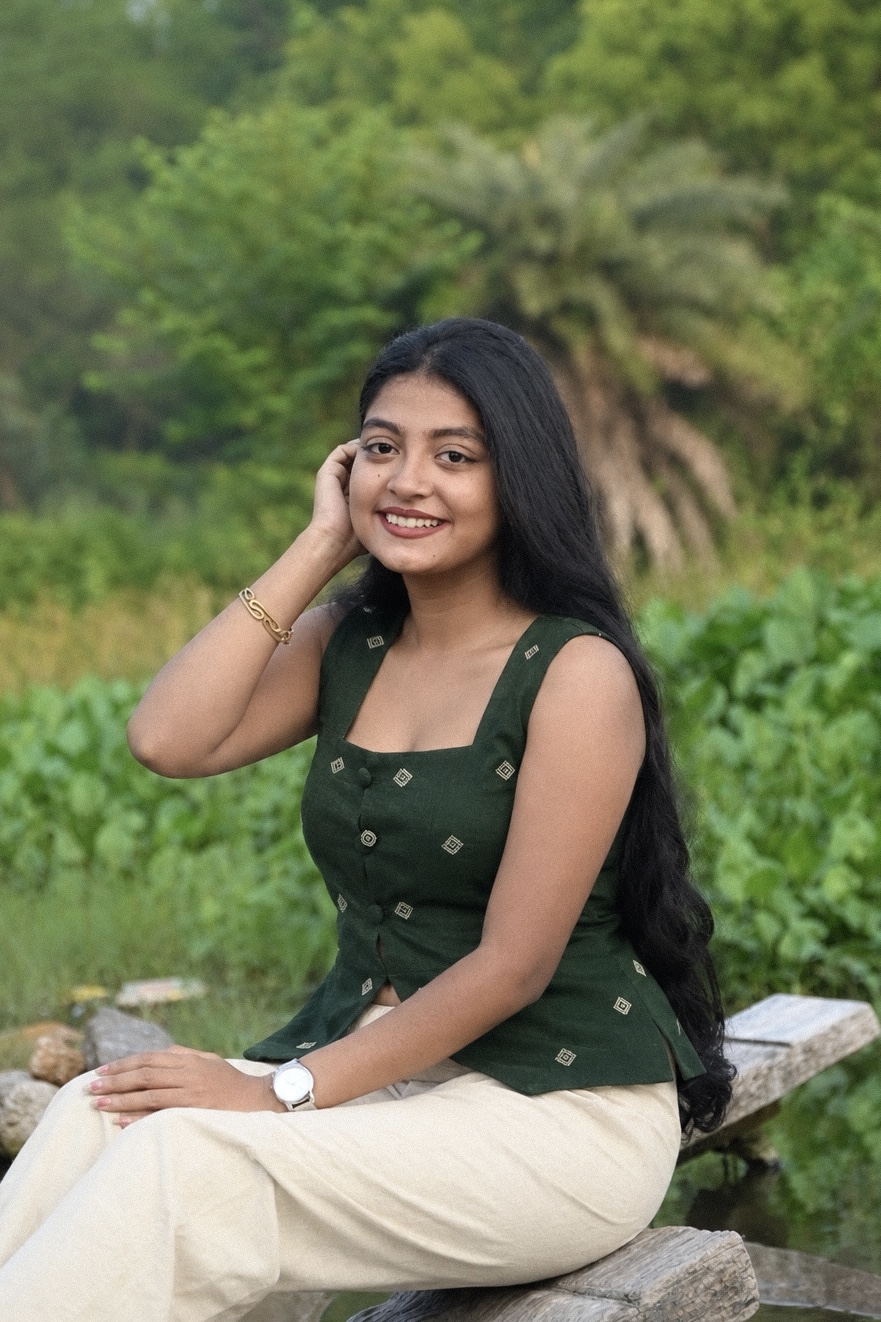 Woman wearing Harshavi’s Aaranya dark green sleeveless top featuring ivory geometric motifs and front buttons, paired with ivory trousers while sitting on a rustic wooden bench in a lush green outdoor