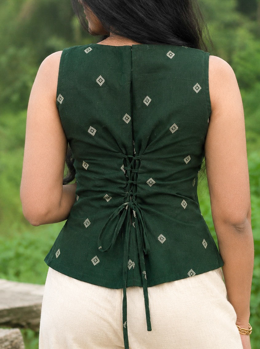 Back view of Harshavi’s Aaranya dark green sleeveless top featuring ivory geometric motifs, a corset-style lace-up back, and adjustable tie detailing, paired with ivory trousers against a lush green o