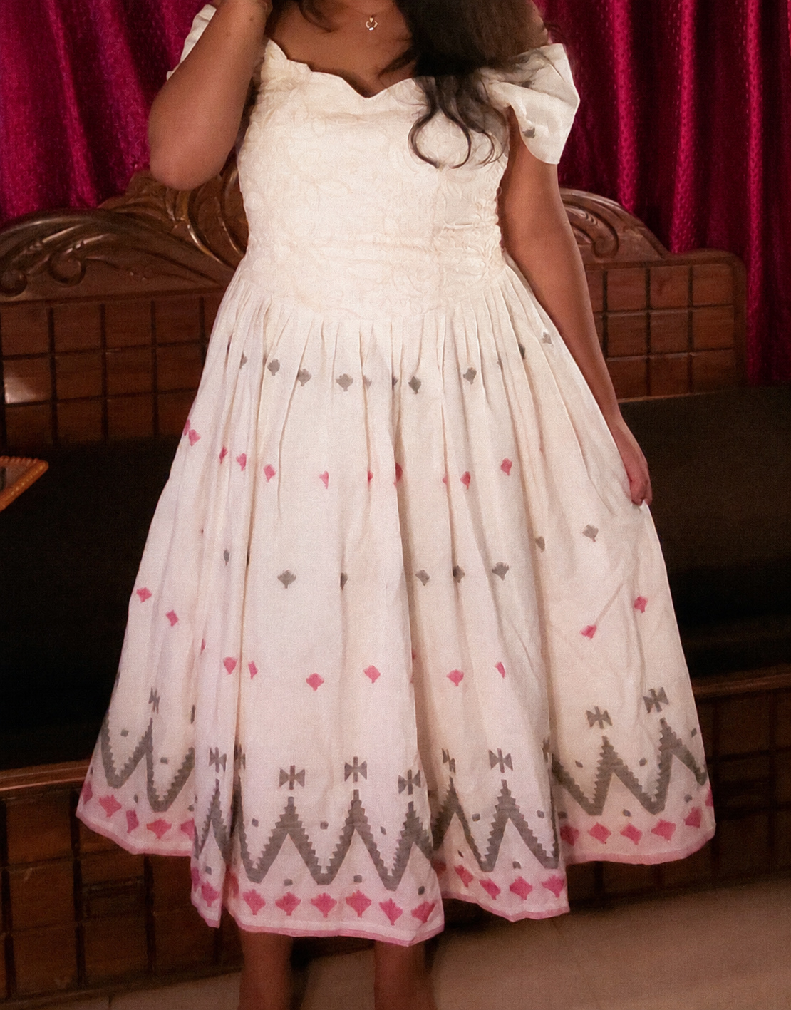 An ivory vintage-inspired dress with puff sleeves, a textured fitted bodice, and a flowing skirt featuring delicate pink and grey traditional geometric motifs, photographed against a rich red velvet