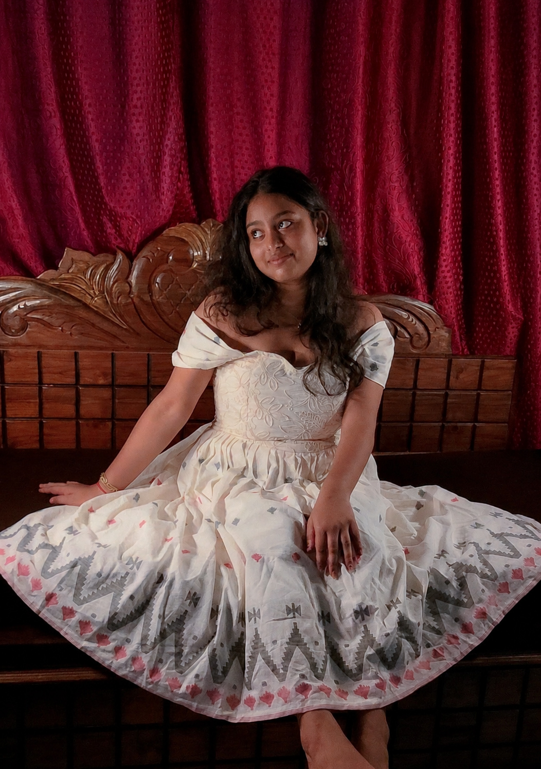 Woman in an ivory embroidered dress sitting on a carved wooden bed against rich burgundy curtains
