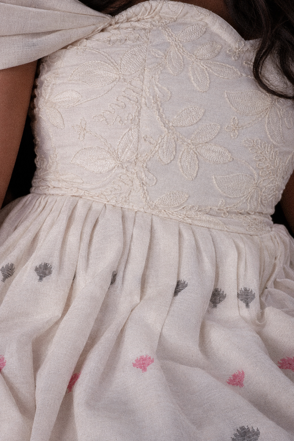 Close-up of an ivory dress featuring intricate floral embroidery, gathered pleats, and scattered dusty-pink and charcoal motifs.