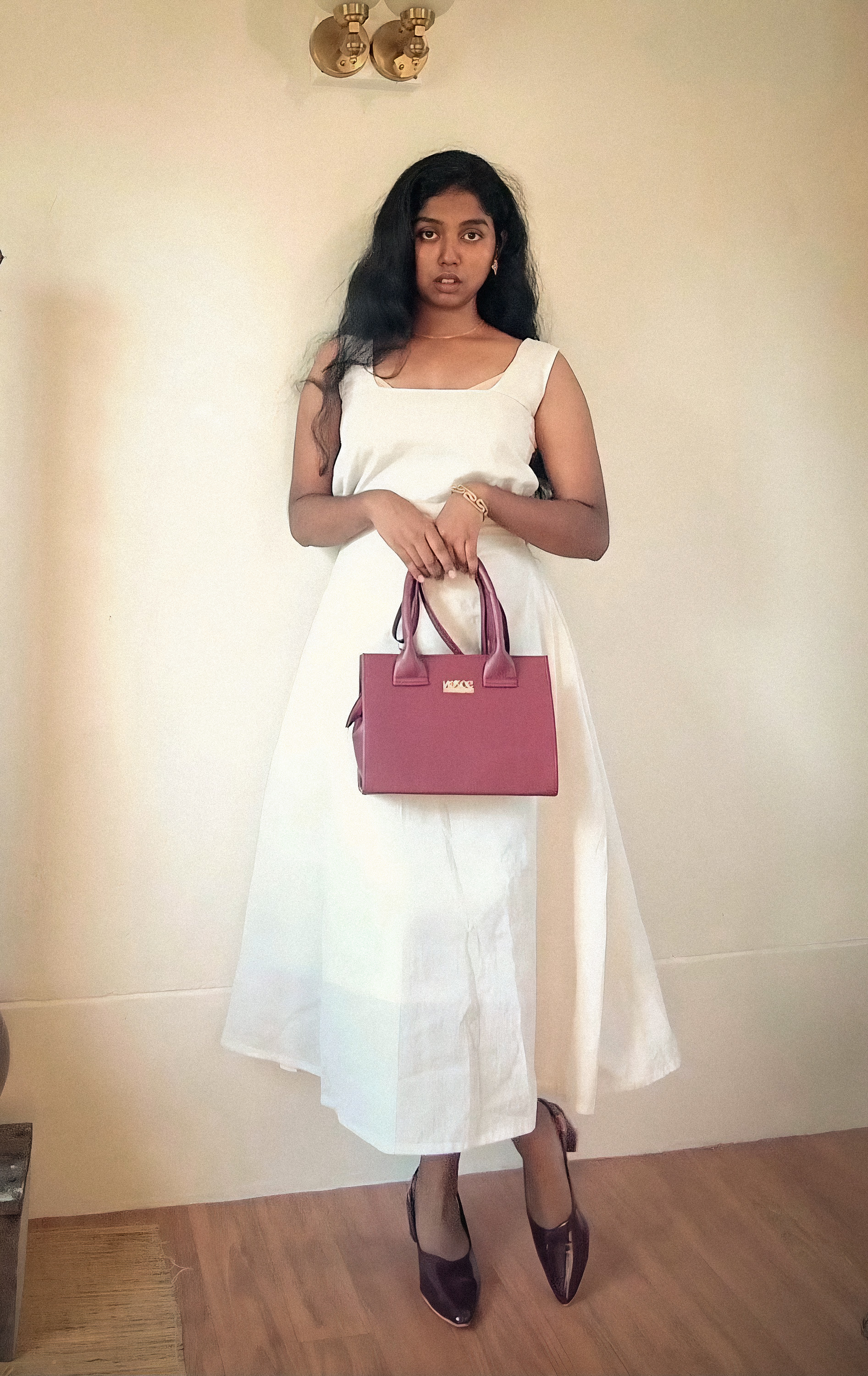 A woman in a white dress standing against a wall while holding a maroon handbag and wearing matching heels.