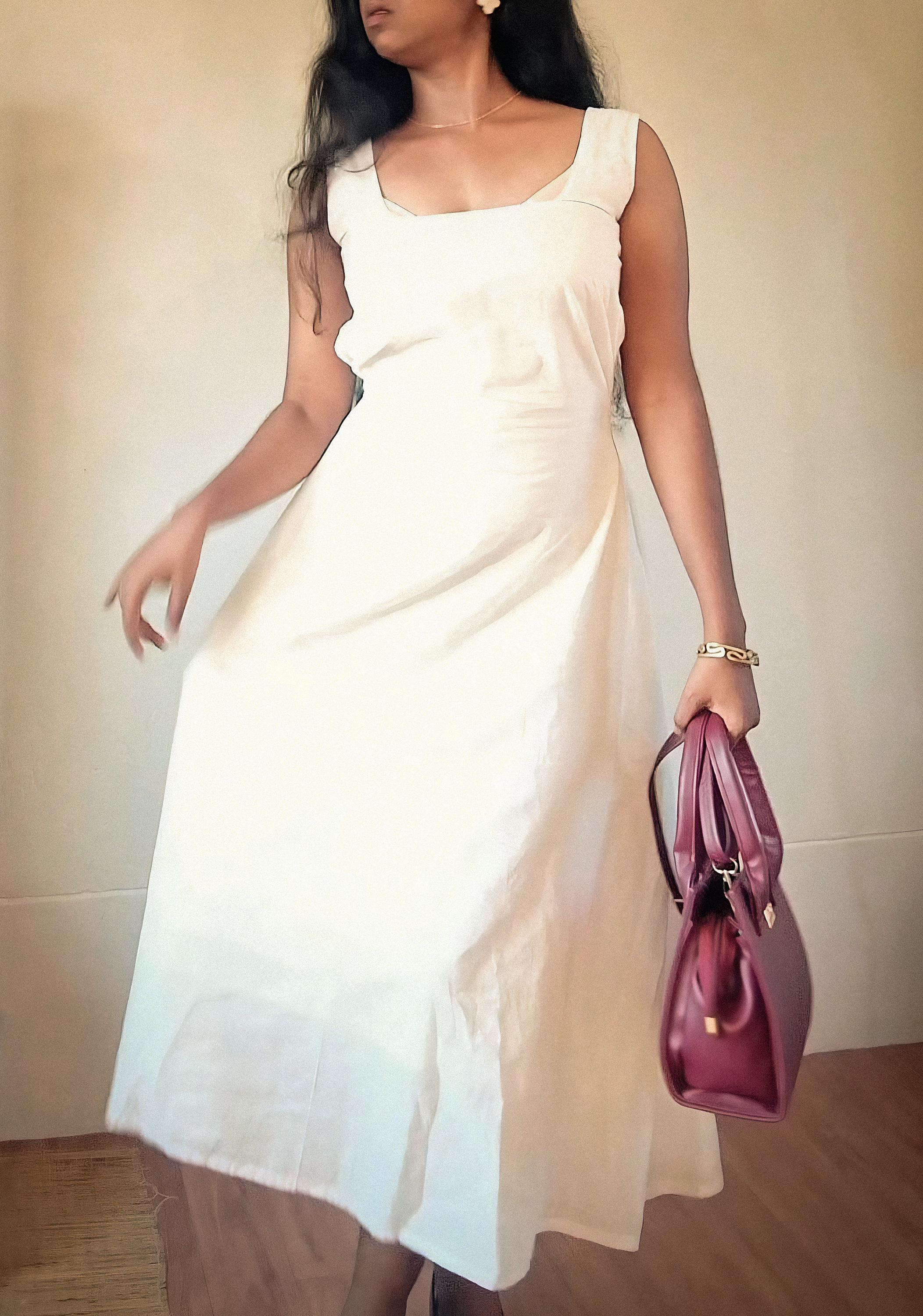 A person wearing a white sleeveless dress and holding a maroon handbag while standing indoors.