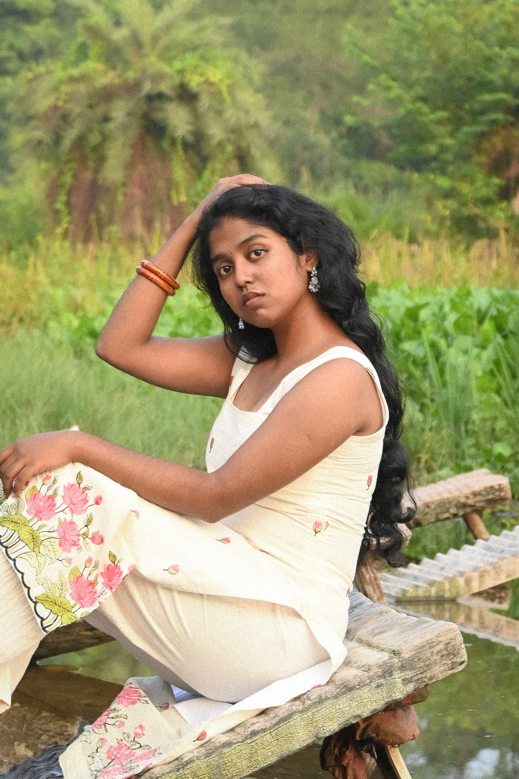 Woman wearing an ivory sleeveless floral kurta with delicate pink bud prints and a pink-and-green floral border, paired with cream trousers, sitting on a rustic wooden log beside a pond with one hand