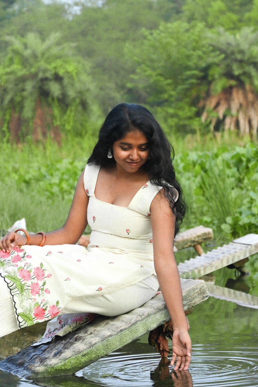 A woman wearing an ivory floral kurta with pink bud prints and a pink-and-green floral hem, paired with cream trousers, sits on a rustic wooden log above a pond, gently touching the water.